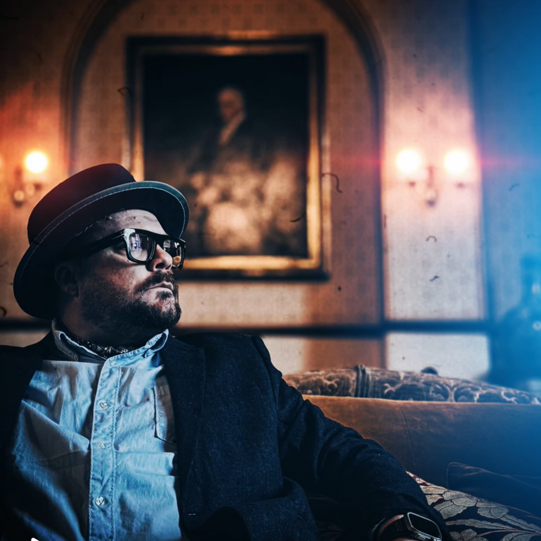 A man wearing a felt hat and glasses, seated in a candlelit room with a portrait on the wall behind him, looking thoughtfully into the distance.